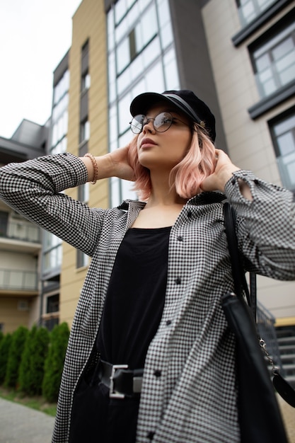 Urban fashion young woman model in fashionable casual clothes and a vintage cap with glasses and a bag walks on the street near the building