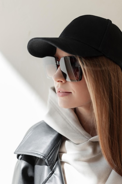 Urban female portrait of beautiful street fashion woman hipster with black cap and sunglasses in trendy leather coat and fashionable hoodie stands near a wall in sunlight on the street