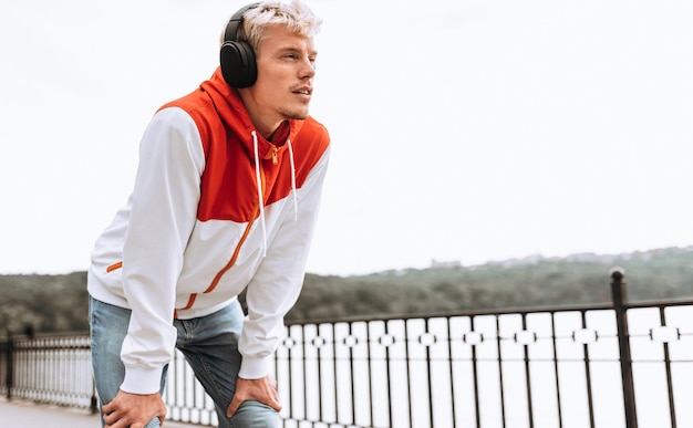 Urban image of handsome young man enjoying weather in city park waiting for his girlfriend while listening to his favorite music from wireless headphones outdoor in the park