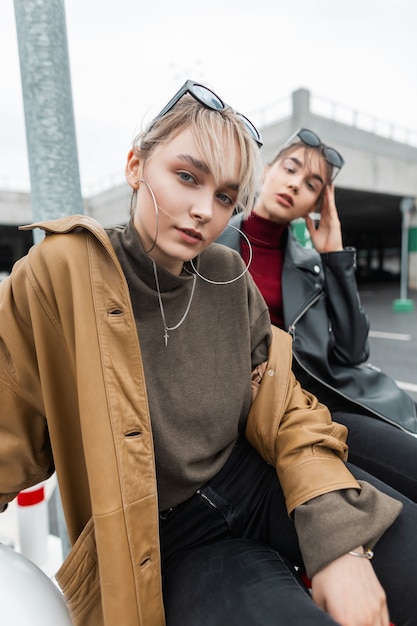 Urban portrait of beautiful young women with leather jacket and sweater in black jeans sits and poses on the street