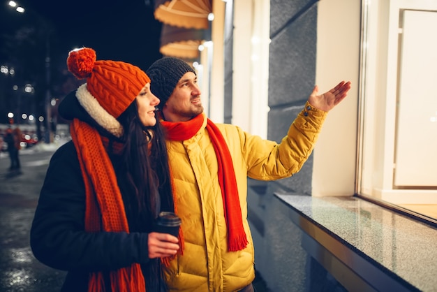 Urban winter evening, love couple looking on showcase. Man and woman having romantic meeting on city street