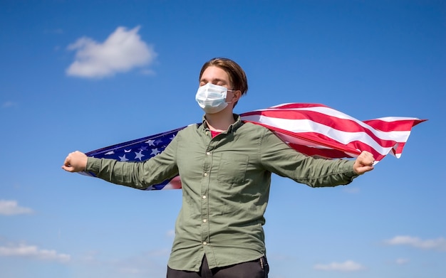 USA flag, a young man in a medical mask holds the USA flag against a blue sky