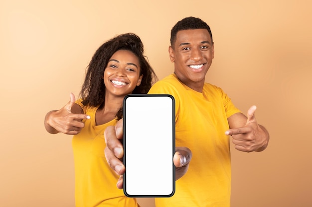 Use this new application Happy black couple pointing at smartphone with blank screen advertising your website mockup