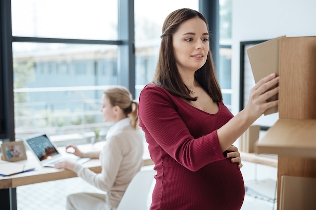 Useful literature. Close up of young pregnant woman standing at the bookshelf while her colleague working with a laptop