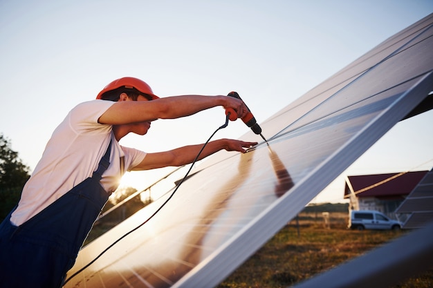 Using cordless screwdriver. Male worker in blue uniform outdoors with solar batteries at sunny day.