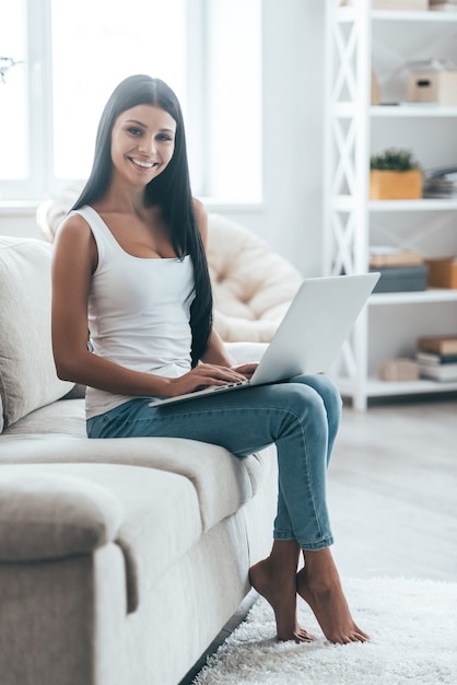 Using modern technologies. Attractive young woman looking at camera and smiling while relaxing on the couch at home