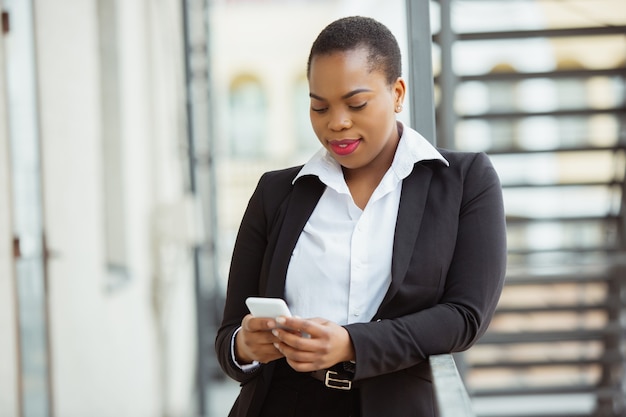 Using smartphone Africanamerican businesswoman in office attire smiling