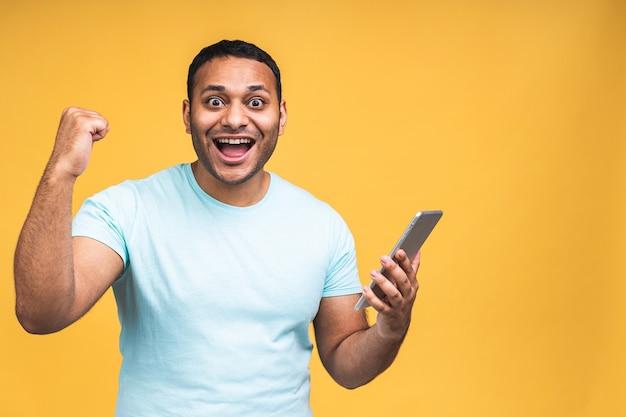 Using tablet. Happy winner! Young handsome african american indian man smiling holding tablet and playing games or using a booking app isolated over yellow background.