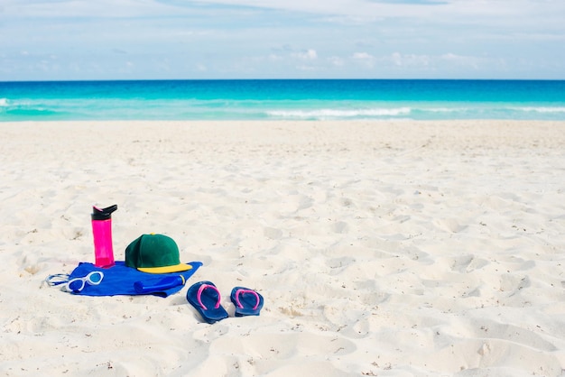 Vacation and summer holidays concept flip flops towel cap bottled water and bottle of sunscreen oil on beach sand