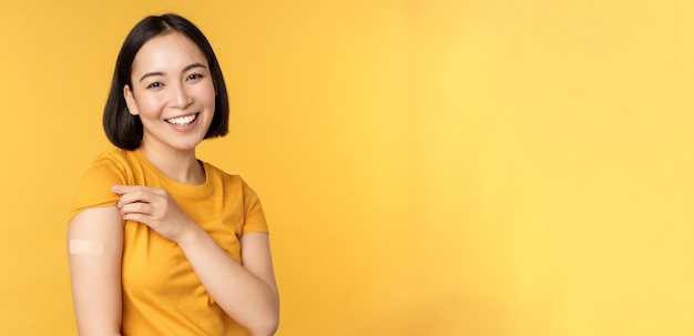 Vaccination and covid19 pandemic concept Happy and healthy asian girl pointing at her shoulder with band aid after vaccinating from coronavirus yellow background