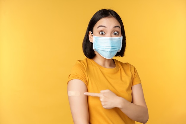 Vaccination from covid and health concept Excited asian woman in medical face mask pointing finger at shoulder with band aid standing over yellow background