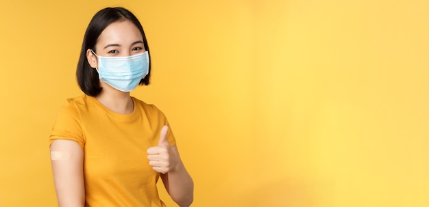 Vaccination from covid and health concept Happy asian girl showing thumbs up wearing medical mask band aid on shoulder got coronavirus vaccine shot yellow background