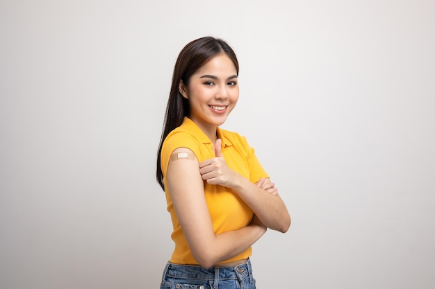 Vaccination. Young beautiful asian woman in yellow shirt getting a vaccine protection the coronavirus. Smiling happy female showing arm with bandage after receiving vaccination. Showing thumbs up.