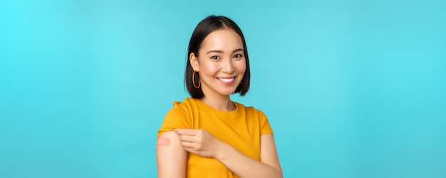 Vaccine campaign from covid19 Young beautiful healthy asian woman showing shoulder with bandaid concept of vaccination standing over blue background