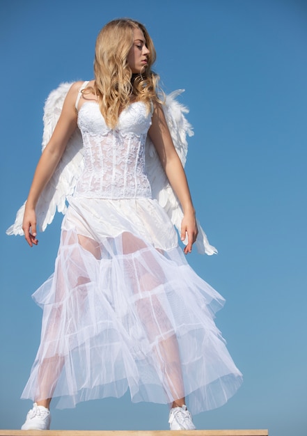 Valentine angel girl with white wings. Womanwith angel wings with bow and arrow against blue sky. Teen angel.