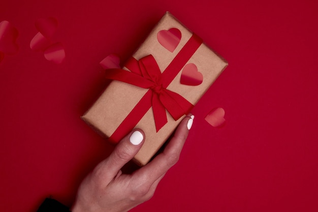 Valentine day composition with copy space. Woman hand holding present or gift box wraped in craft paper with ribbon bow and red hearts confetti. Close up, top view, copy space