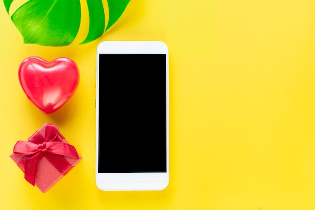 Valentine Day online Shopping. Seasonal Holiday sale. Smartphone, red heart, gift, monstera on yellow trend background. Romantic shopping for gerls, womens day, buying gifts Valentine's Day lovers