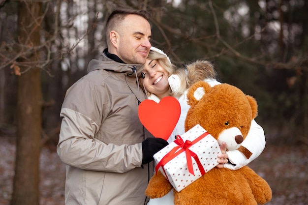 Valentine's Day celebration at happy couple in winter forest. Smiling woman in white jacket is holding gift box and Teddy bear. Man embraces woman and holds red heart in his hands. Surprises.