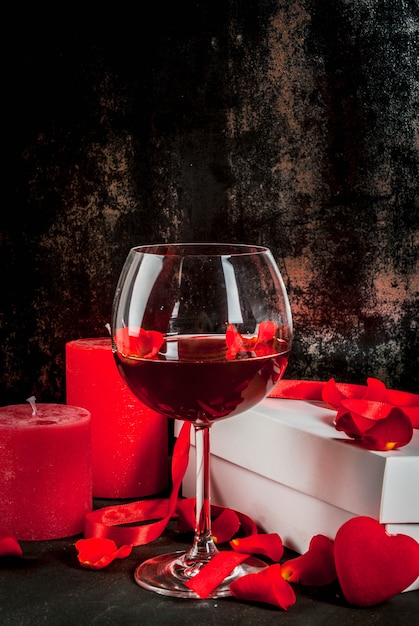 Valentine's day concept, white wrapped gift box with red ribbon, rose flower petals, red wine glass, with red candle, on dark stone background, copy space