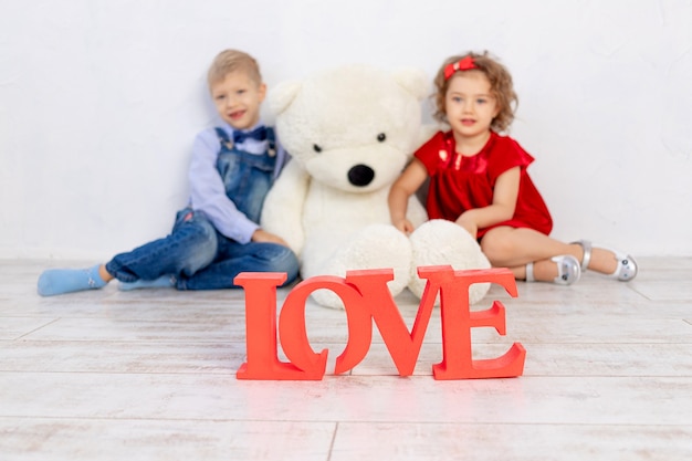 Valentine's day kids. Boy and girl sit with a big teddy bear, the inscription love in focus