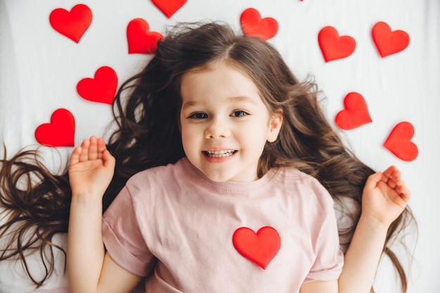 Valentine's Day a little girl lying at home on a white bed among hearts smiling and laughing with happiness congratulating on the holiday