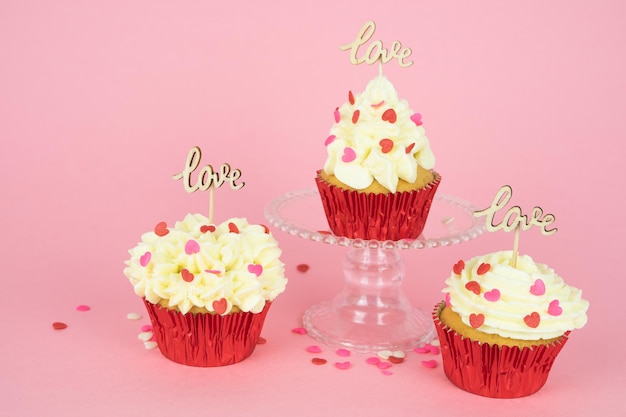 Valentine's Day. Three cupcakes with cream and red topping decoration with a wooden sign with the word LOVE on a pink background. Copy space.