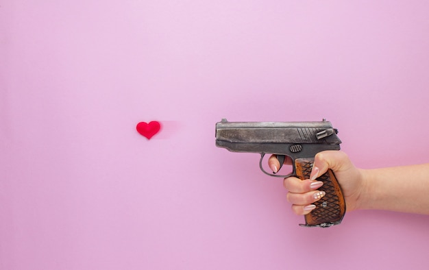 Valentine's Day . Womans hand holding a gun and shooting with heart on pink background.