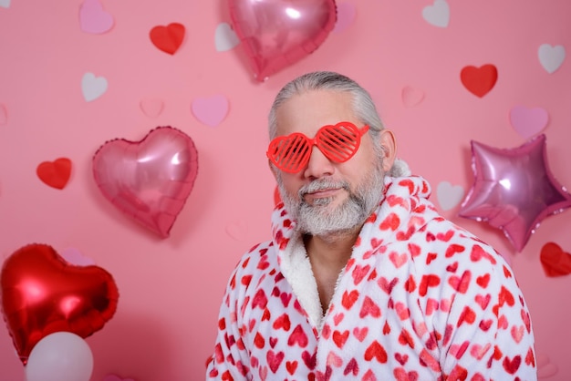 Valentine39s Day portrait of grayhaired man wearing heartshaped glasses and bathrobe with red hearts