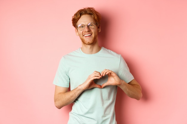 Valentines day concept. Handsome redhead man in glasses, showing heart sign and say I love you, standing over pink background.