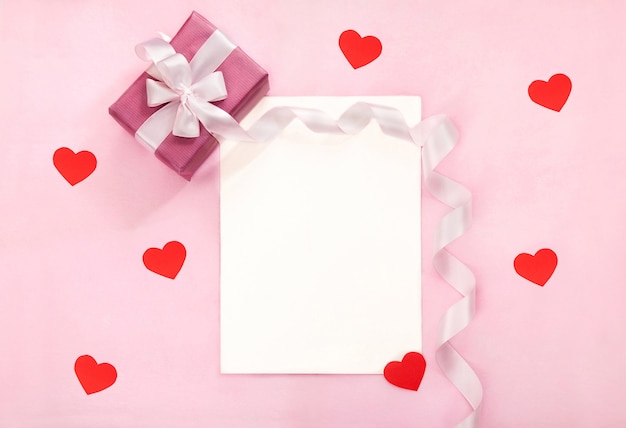 Valentines day greeting card with pink gift box, white bow, long curved ribbon and paper red hearts