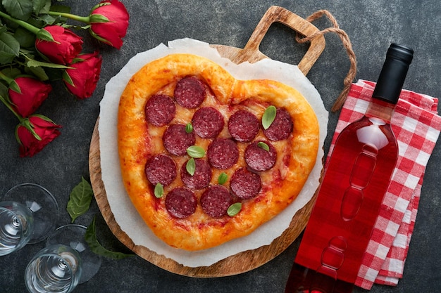 Valentines day heart shaped pizza with mozzarella pepperoni and basil wine bottle two wineglass gift box on black background Idea for romantic dinner Valentines day Top view Mock up