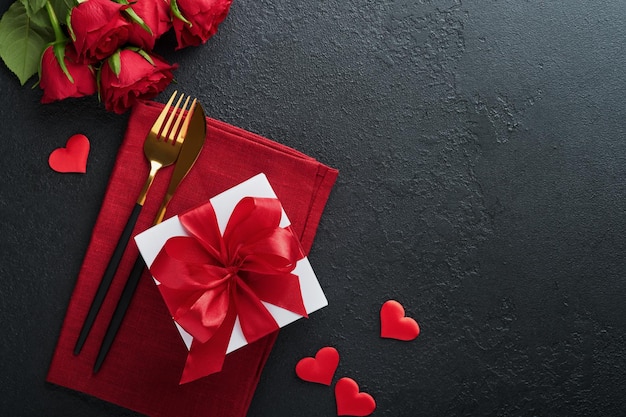 Valentines day romantic table setting Empty closeup of red roses wine candles dinner black plate knife fork and decorative silk hearts on black background Holiday concept Copy space Top view