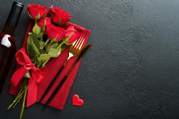 Valentines day romantic table setting Empty closeup of red roses wine candles dinner black plate knife fork and decorative silk hearts on black background Holiday concept Copy space Top view