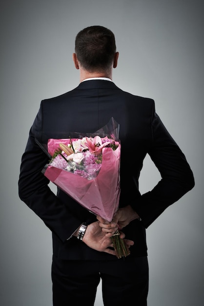 Valentines day surprise and flowers of a man in a suit with love present behind back Flower gift suit and male model in a isolated gray background studio feeling romantic for a formal date