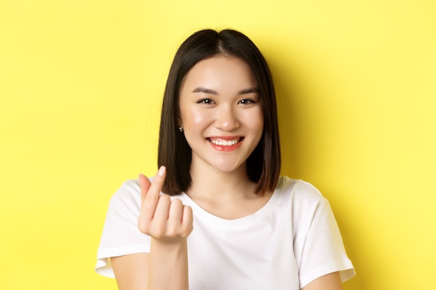 Valentines day and women concept. Close up of pretty asian girl in white t-shirt, smiling and showing finger heart, standing over yellow background.