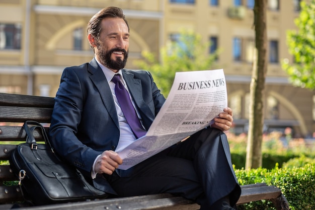 Valid information. Positive handsome businessman reading a newspaper while sitting on the bench