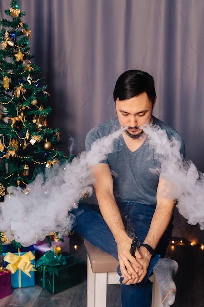 Vaper exhales steam. The guy sits near the Christmas tree and smokes an electronic cigarette.