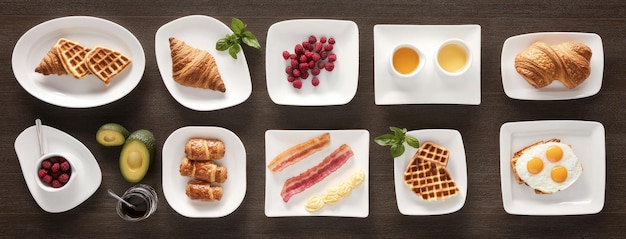 A variety of breakfast dishes including eggs bacon croissants waffles and fruit arranged on white plates over a dark wooden table