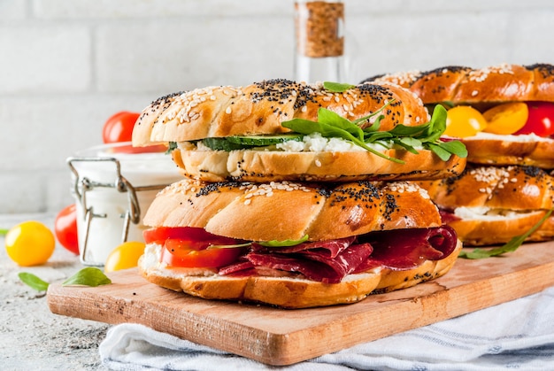 Variety of homemade bagels sandwiches with sesame and poppy seeds, cream cheese,  ham, radish, arugula, cherry tomatoes, cucumbers, white grey textured surface 