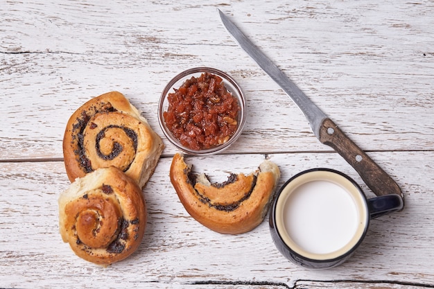 Variety of homemade puff pastry buns cinnamon served with milk cup, jam, butter as breakfast over white plank wooden background.