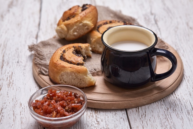 Variety of homemade puff pastry buns cinnamon served with milk cup, jam, butter as breakfast over white plank wooden table. Flat lay, space