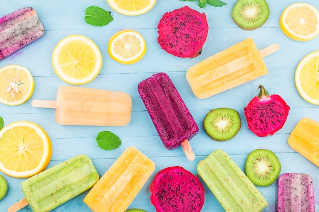 Various fruit popsicles are placed on the blue wooden board background, kiwi popsicles, orange popsicles, dragon fruit popsicles, cantaloupe popsicle