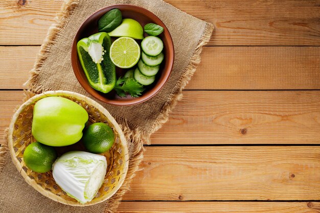 Various green fruits and vegetables on a wooden table. Chopped green apple, bell pepper, cucumber, cabbage and lime in a clay bowl. Top view