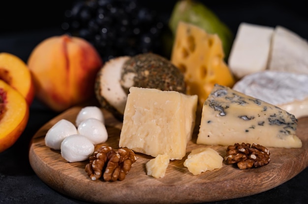 Various types of cheese on a dark background. Nearby are peaches, grapes, nuts. An exquisite delicacy.