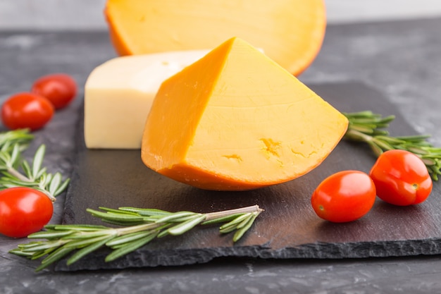 Various types of cheese with rosemary and tomatoes on black slate board on a black concrete surface. Side view, close up.