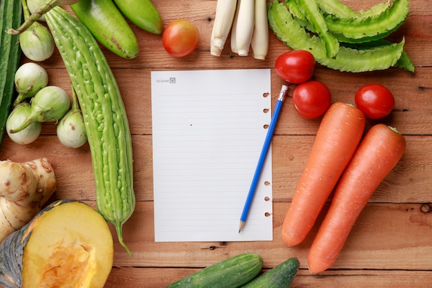  Various vegetables with blank note page and pencil on wooden background. Top view 