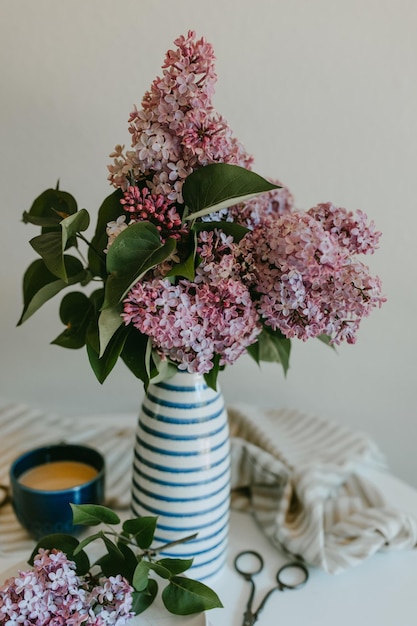 A vase of lilacs sits on a table with a cup of coffee