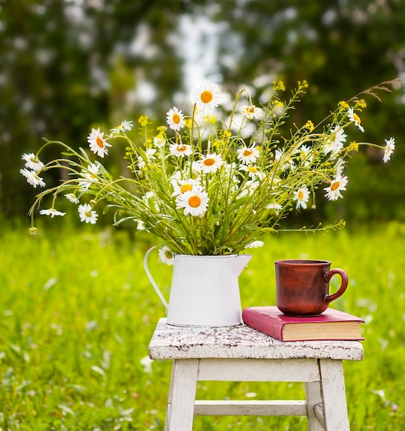 a vase with daisies, a book and a mug on a stool outdoors