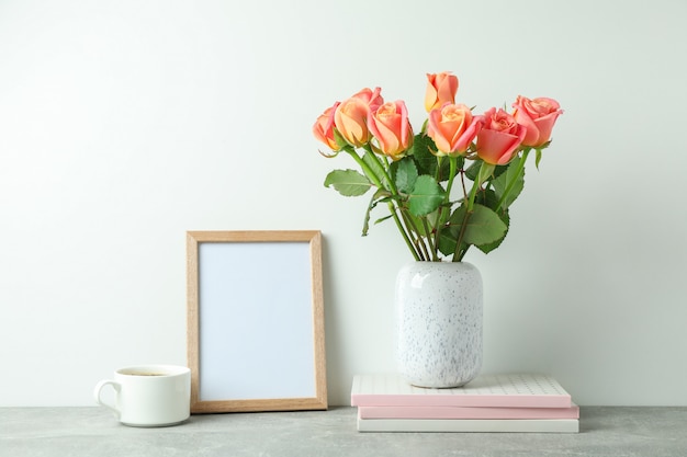 Photo vase with pink roses, copybooks, empty frame, cup of coffee on grey table