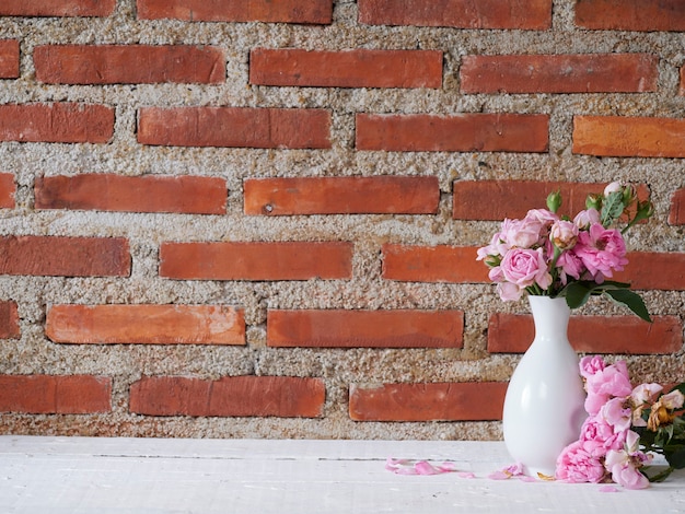 Vase with pink roses on white table near brick wall